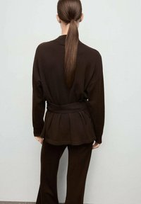Top de punto marrón con cintura ceñida y cinturón, que presenta una parte trasera plisada y mangas largas, combinado con pantalones marrones a juego. Textura suave, diseño sencillo.
