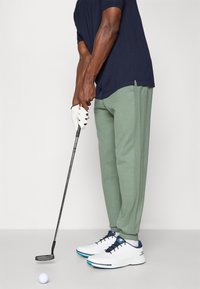 Golf apģērbs, kas sastāv no tumši zilā polo krekla, gaiši zaļiem sporta biksēm un baltām kedām ar zilo akcentu. Tur rokā golfu nūju blakus baltai bumbai.