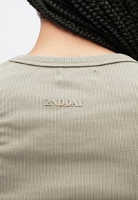 Tricou verde măsliniu cu dungi, decolteu rotund, cu un logo subtil "2NDDAY" cusut pe spate, prim-plan al texturii țesăturii.