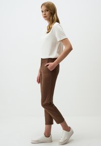 Top bianco a maniche corte abbinato a pantaloni slim-fit marroni, con tasche laterali. Indossato con sneakers bianche, su uno sfondo chiaro e semplice.