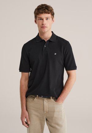 Noor mees, kes kannab musta lühikese varrukaga polosärki ja beeže pükse, seisab ühe käega taskus ühevärvilise tausta ees.