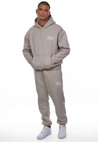 Beige hoodie og matchende sweatpants sæt lavet af blødt stof. Hoodien har en frontlomme og grafisk tekst på brystet og bukserne. Hvide sneakers.