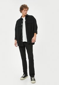 Camisa negra de gran tamaño con dos bolsillos frontales, superpuesta a una camiseta blanca, combinada con pantalones negros ajustados y zapatillas negras con suelas blancas.