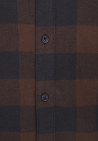 Camisa de franela a cuadros que presenta colores gris oscuro y marrón, con un cierre de botones en la parte delantera y un tejido texturizado. Los botones de plástico negro detallan el cierre.