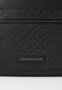 Pouch in materiale sintetico nero con texture, caratterizzato da schemi geometrici in rilievo. Logo del marchio stampato su un'etichetta rettangolare. Chiusura con zip.