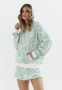 Zestaw bluzy z kapturem i szortów w jasnym zielonym kolorze z białym spiralnym wzorem, wykonany z miękkiej tkaniny, z przednią kieszenią i elastycznymi mankietami.