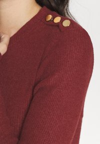 Bordowy dzianinowy sweter o prążkowanej fakturze, z trzema metalicznymi guzikami na ramieniu. Miękki i dopasowany krój.