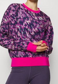 Langärmliger Nike Sweatshirt aus dunkel lila Stoff mit abstrakten pinken und silbernen Mustern sowie leuchtend pinken Bündchen und Kragen.