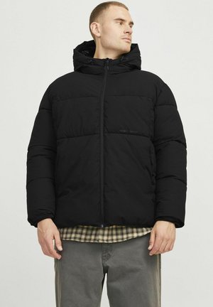 Schwarze Daunenjacke mit Kapuze, ausgestattet mit einem gesteppten Design, Frontreißverschluss und Logo auf der Brust. Getragen über einem karierten Hemd und grauen Hosen.