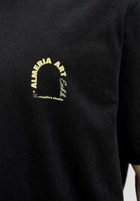 Čierne bavlnené tričko s krátkymi rukávmi s žltým potlačeným vzorom, ktorý obsahuje "ALMERIA ART" a "Kreatívne štúdio."
