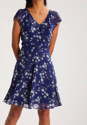 Donna che indossa un vestito floreale blu navy con maniche corte, scollo a V e una sottile cintura in vita, in piedi davanti a uno sfondo semplice.
