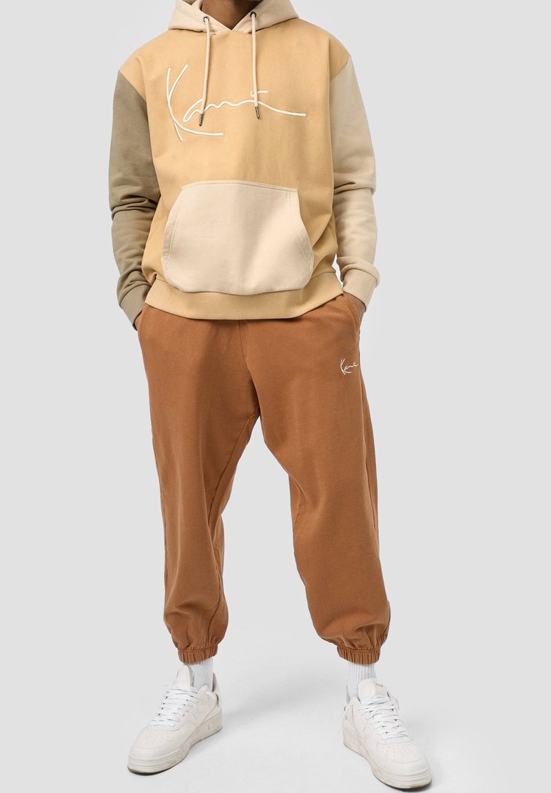 Beige huvtröja med en framficka och vitt logotyp, kombinerad med bruna joggerbyxor med ankelmanschetter, kompletterad med vita sneakers.