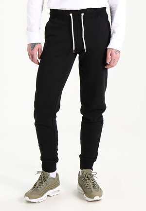 Mężczyzna ubrany w czarne spodnie dresowe z białymi sznurkami, białe skarpetki i oliwkowozielone sneakersy stoi na białym tle.