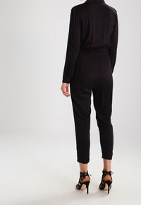 Combinaison noire avec une taille ajustée, des manches longues et des poignets. Stylisée avec des sandales à talons hauts noires à lanières. Tissu lisse avec une ceinture élastique.