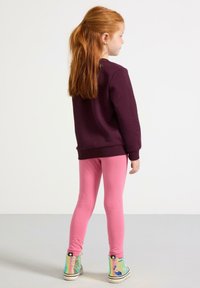Bordeaux sweatshirt med ribbet manchetter, parret med lyserøde leggings. Skinnende, multifarvede ankelstøvler fuldender outfittet. Simpelt, ensfarvet baggrund.