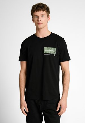 Jonge man met krullend haar die een zwarte T-shirt draagt met een klein groen grafisch patchje op de linkerborst, staand tegen een effen witte achtergrond.