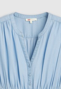 Světle modrá chambray blůza s výstřihem do V, nařaseným pasem a knoflíkovou légou se pěti knoflíky. Měkká bavlněná tkanina, nařasený detail.