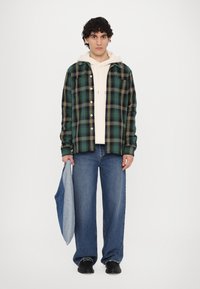 Grønn og gul rutete skjorte med knapper, lagdelt over en beige hettegenser. Løse blå denimjeans og svarte sko. Grå-blå veske holdt i hånden.
