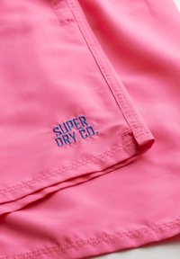 Tessuto rosa con una superficie liscia, caratterizzato dalla scritta "SUPER DRY CO." ricamata in blu. Il design include un orlo cucito e linee pulite.
