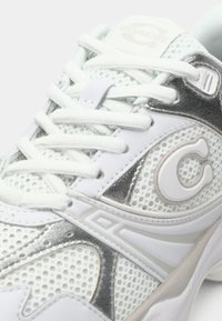 Witte sportieve sneaker met meshpanelen, zilveren accenten, textuurdetails en platte witte veters. Coach-logo nabij de tong.