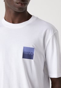 White cotton t-shirt with a crew neck, featuring a rectangular blue logo patch that includes the word "HUGO" in a darker shade.