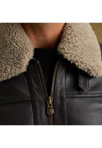 Čierna kožená bunda s krémovým, nadýchaným umelým ovčím golierom. Obsahuje zlatý zips a horizontálne švy. Detailný pohľad na výstrih.