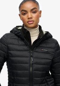 Giacca a piumino nera con cappuccio, caratterizzata da una finitura testurizzata e chiusura frontale con cerniera. Presenta un interno verde a contrasto e un logo Superdry.