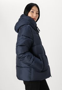 Ženska v mornarsko modri jakni z zadrgo in kapuco, z rokami v žepih, ki se obrača čez ramo proti enobarvni svetli podlagi.