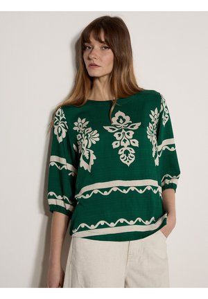 Femme portant un chemisier vert à motifs floraux blancs et manches trois-quarts, associé à un pantalon beige clair, debout contre un mur uni.