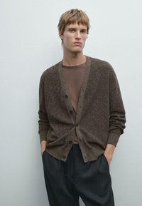 Brown textured cardigan with button closure, ribbed cuffs, and pockets, layered over a brown T-shirt, paired with dark tapered pants.
