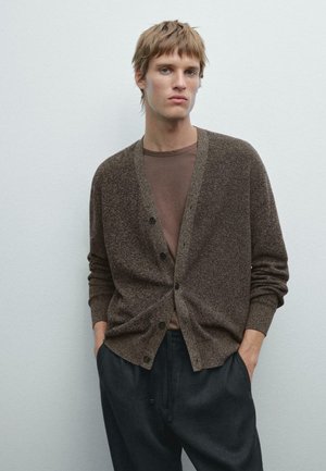 Brown textured cardigan with button closure, ribbed cuffs, and pockets, layered over a brown T-shirt, paired with dark tapered pants.