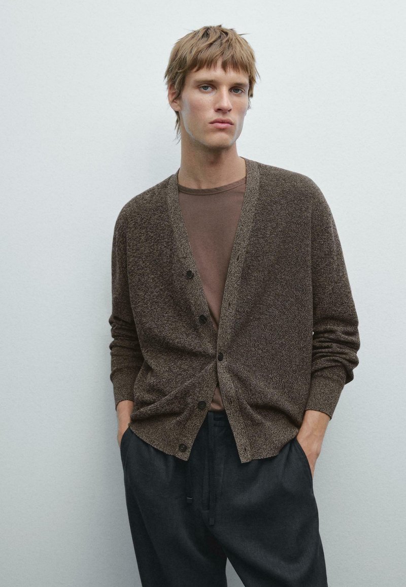 Brown textured cardigan with button closure, ribbed cuffs, and pockets, layered over a brown T-shirt, paired with dark tapered pants.