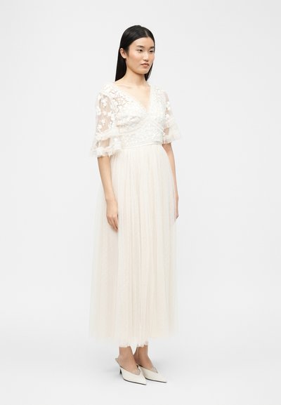 Γυναίκα με μακριά σκούρα μαλλιά που φοράει λευκό φόρεμα V-λαιμόκοψη με δαντέλα και λουλούδια, διαφανή φουσκωτά μανίκια και λευκές μυτερές γόβες, στέκεται μπροστά σε απλό φόντο.