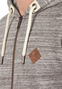 Sudadera gris con cremallera y tela texturizada, con cordones beige, parche de logo de cuero marrón y herrajes de cremallera de color cobre.