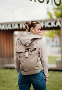 Béžový batoh s rolovacím vrškem a puntíkovaným detailem, s přední kapsou na zip a nastavitelnými popruhy, v kombinaci s lehkou bundou a modrými džíny.