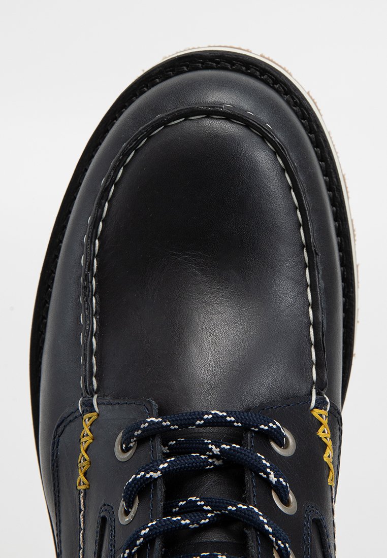 Botte en cuir noir avec bout arrondi, surpiqûres blanches et lacets navy. Présente des accents jaunes près des œillets pour plus de détails.