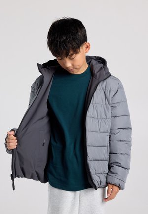 Jongen draagt een grijze donzen jas, die hij openritst om de donkere voering en een donkergroen shirt eronder te laten zien.