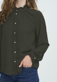 Femme portant une blouse noire à manches longues avec un col et des poignets volants, la main dans la poche d'un jean en denim bleu.