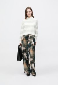 Sweter w jasnoszare i białe paski, zestawiony z luźnymi, ciemnymi spodniami w kwiatowy wzór. Modelka trzyma czarną torbę typu tote, stoi na neutralnym tle.