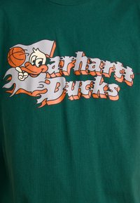 T-shirt en coton vert avec le texte "Carhartt Ducks" en orange et blanc, accompagné d'un graphique de canard cartoon et de basketball. Coupe classique, manches courtes.