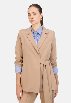 Blazer beige su misura con vita sottolineata da una cintura, abbinato a una camicia a righe blu e bianche con colletto, polsini abbottonati in vista. Texture liscia.