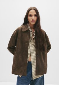 Hnědá sueded bunda s uvolněným střihem, velkým límcem a dvěma knoflíky. Nositá přes kostkovanou košili, která ukazuje texturovanou tkaninu a oversized design.