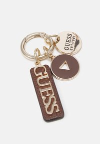 Nøglekæde med brun tekstureret tag med gyldent "GUESS" bogstaver, gyldent cirkulært charm og trekantet brune detalje, alle fastgjort til en metalring.