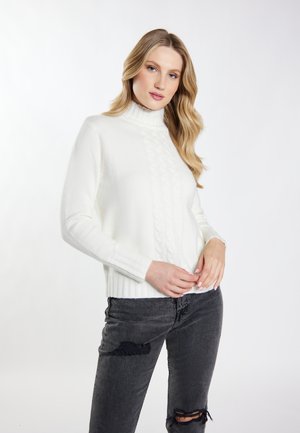Blonde vrouw met losse golven draagt een witte coltrui kabelgebreide trui en zwarte gescheurde spijkerbroek, staand tegen een effen witte achtergrond.