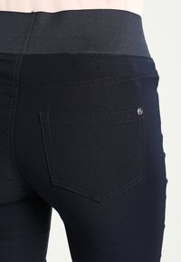 Tmavě modré kalhoty s širokým elastickým pasem, s zadní kapsou a malým knoflíkovým detailem, vyrobené z hladkého a pružného materiálu.