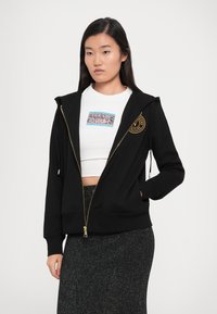 Black zip-up hoodie with gold zipper and embroidered logo on the chest, worn over a white cropped T-shirt with a colorful graphic.