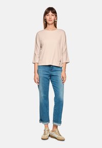 Light beige long-sleeve top with a rounded neckline and side pocket, paired with light blue cuffed jeans and beige sneakers.