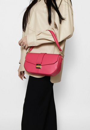 Pink faux leather handbag with a flap closure, gold accent hardware, and a detachable shoulder strap, held at the side against a neutral background.