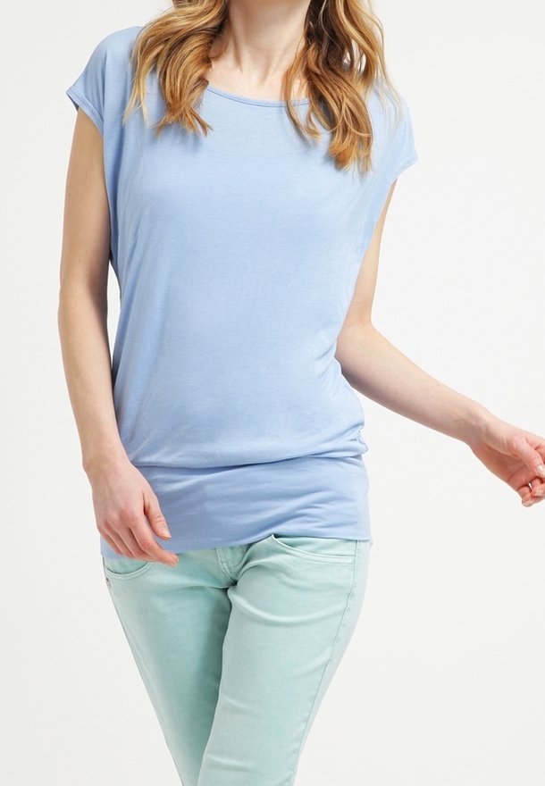 Femme portant un haut à manches courtes bleu clair et un pantalon vert menthe, debout devant un fond blanc uni.