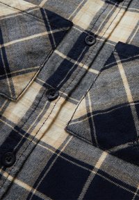 Dettaglio ravvicinato di una camicia a quadri beige e blu navy con chiusura a bottoni e due tasche sul petto con paramontura.